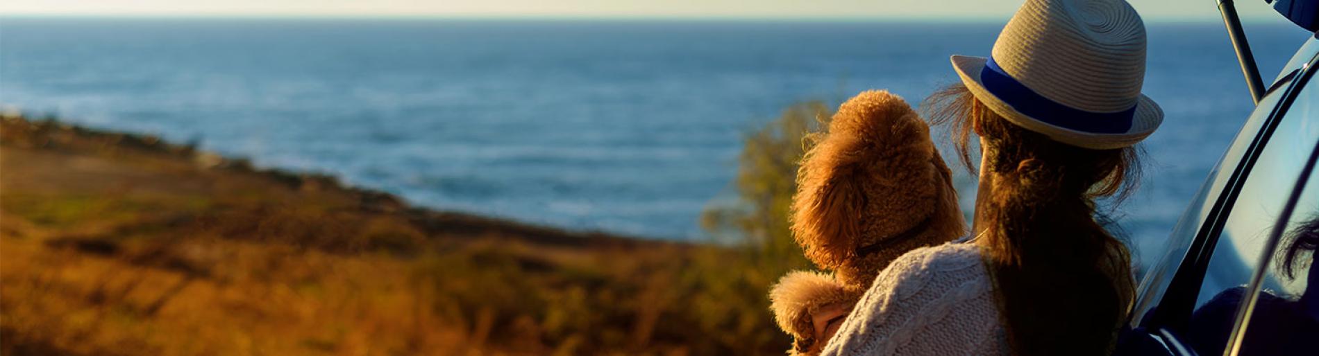 A woman holding her dog while leaning on her car looking at an ocean view